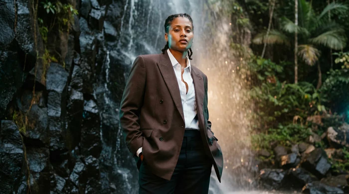 Femme en blazer brun devant une cascade de forêt tropicale martiniquaise, posture calme et assurée — l'exigence professionnelle dans un décor de Martinique, l'esprit Byss Group